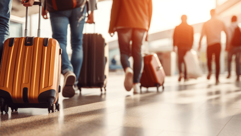 Travelers with rolling suitcases in a sunlit airport terminal, focus on a yellow suitcase.