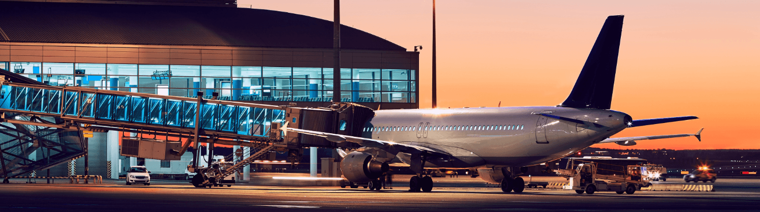 Airplane at a lit terminal during sunset, jet bridge attached, airport apron under a colorful evening sky.
