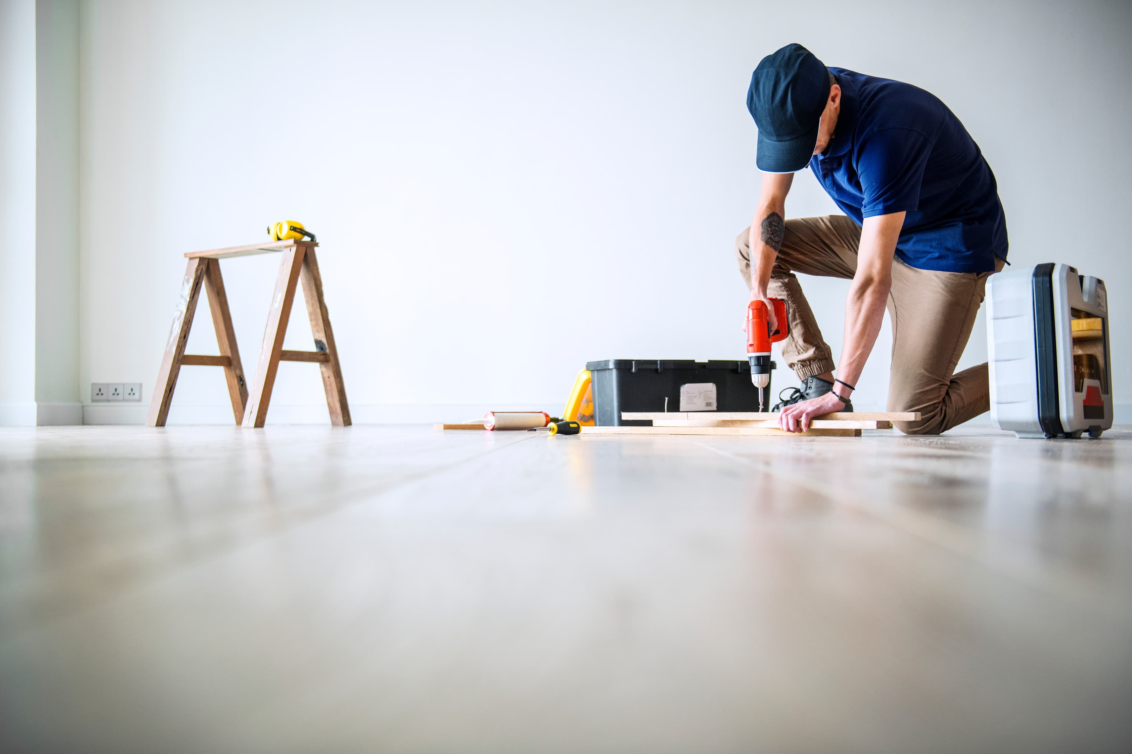 Handyman Service: Man kneeling on wood floor, drilling hole, toolbox and ladder in background.