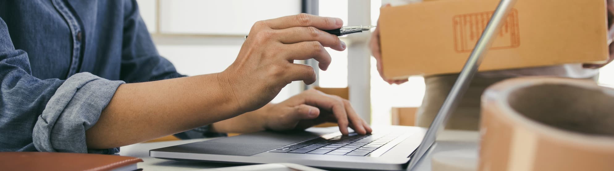 A coordinator working on a laptop next to a moving box and packing tape – symbol image for the strategic procurement of moving and relocation services in B2B procurement.