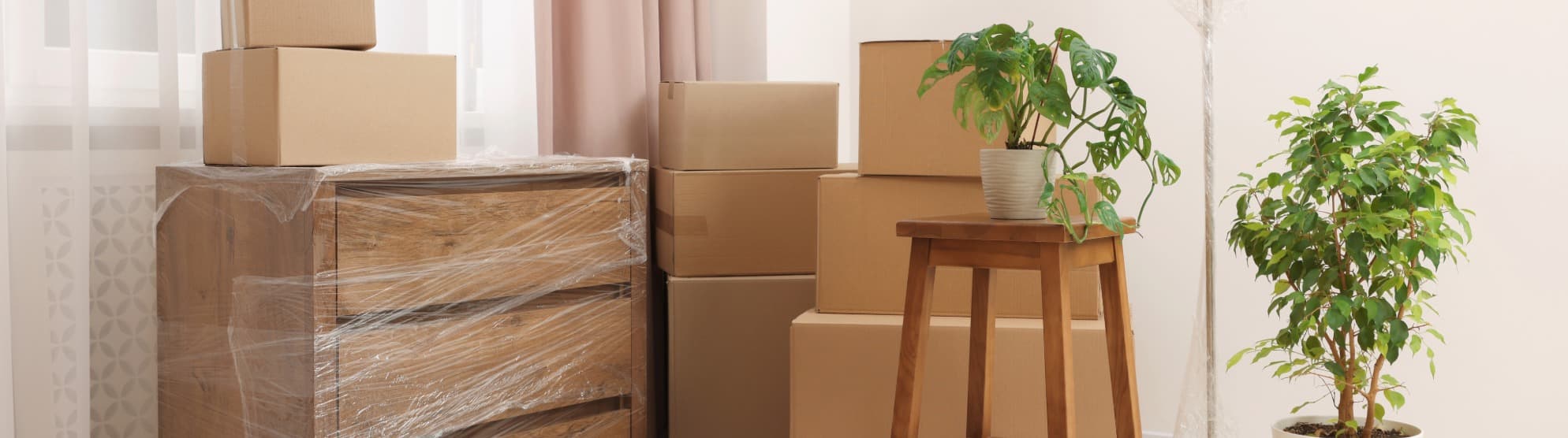 A wooden dresser wrapped in protective stretch film next to stacked moving boxes in a bright apartment, ready for small volume transport with DACHSER & KOLB.