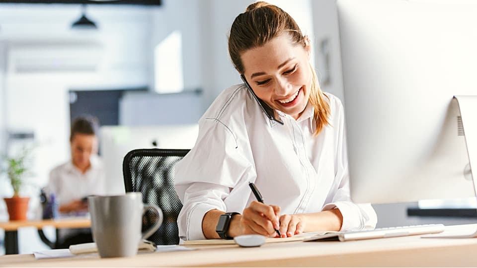 A smiling HR professional speaks on the phone at her computer and notes key budget reporting data.