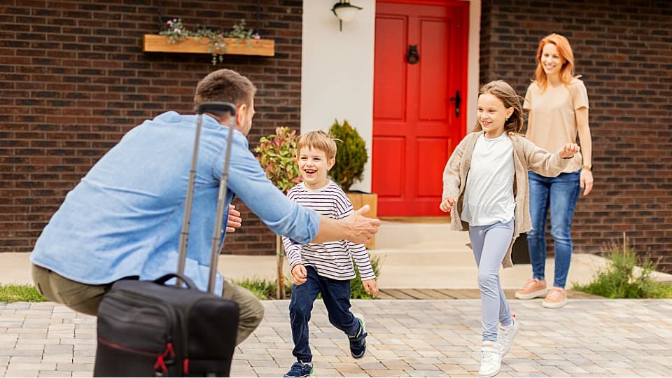 Children happily run toward their father with a suitcase in front of the new home while the mother smiles in the background.