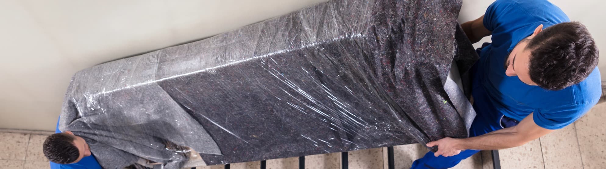 Two movers carefully carry a sofa wrapped in blanket and film through a stairwell.