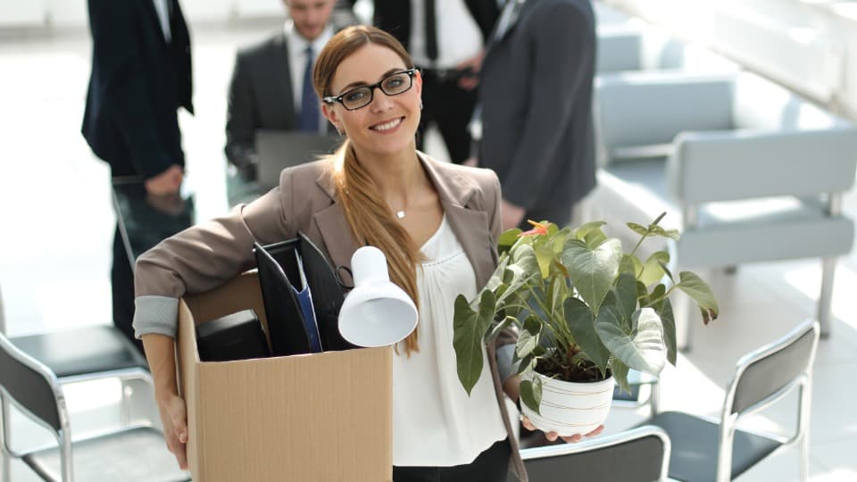 Smiling employee carrying a moving box and a plant to a new workstation.