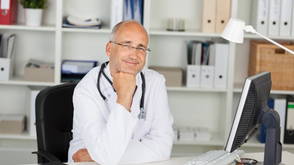 A doctor from Luxembourg is sitting at his desk and smiling at the camera