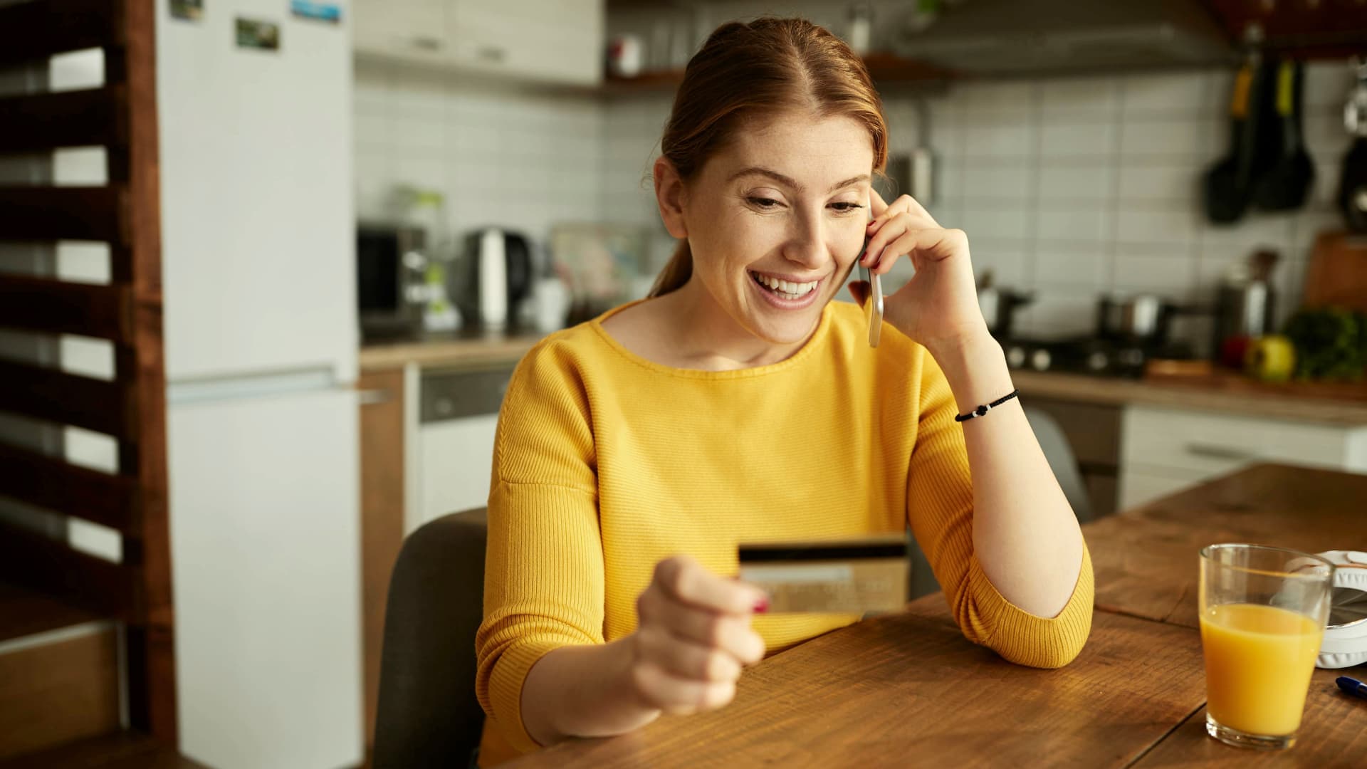 Woman on phone holding credit card at kitchen table, flexible payment for moving on a budget