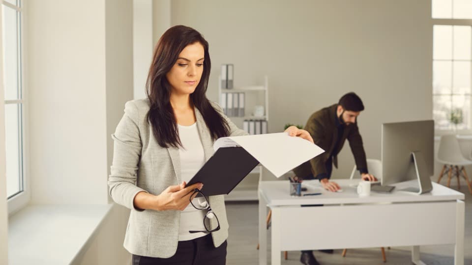 A facility manager reviews office restructuring documents in a bright office while a colleague works at a desk in the background – facilities logistics for facility management.