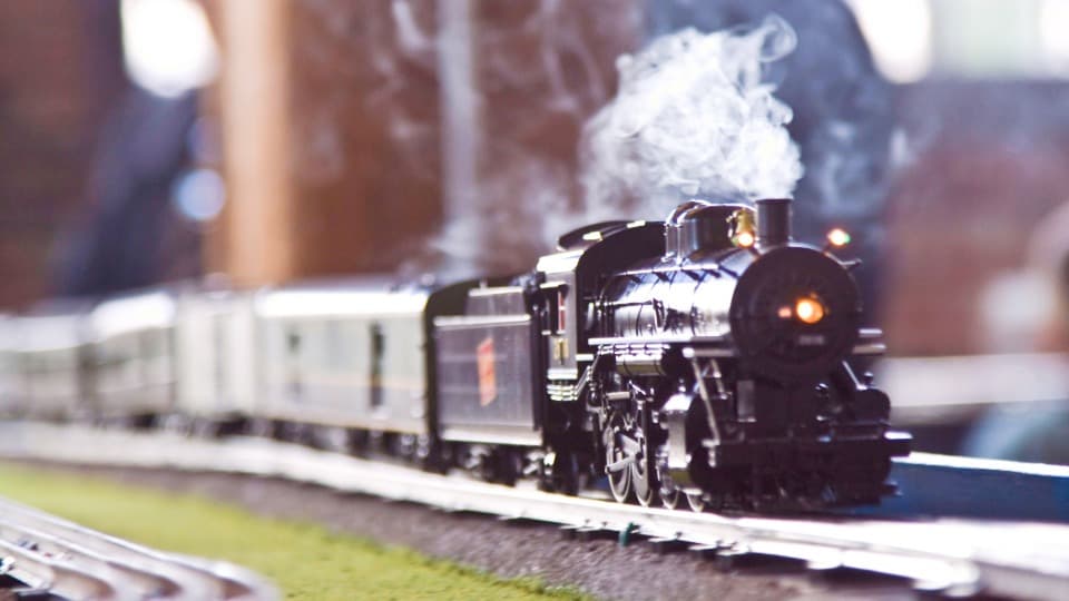 Eine Modelleisenbahn-Dampflokomotive fährt mit Dampf auf den Schienen einer Anlage, deren Transportweg vor dem Umzug mit DACHSER & KOLB genau geprüft werden muss.