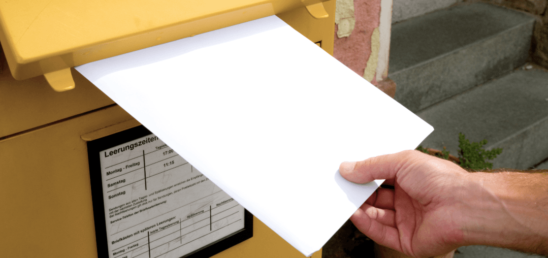 Hand posts a letter into a yellow mailbox – symbolic image for submitting a Nachsendeauftrag (mail forwarding request).