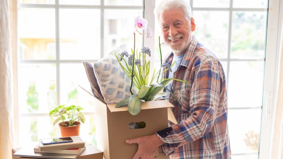 Ein glücklicher älterer Mann trägt beim Seniorenumzug mit DACHSER & KOLB einen Umzugskarton mit einer Orchidee.