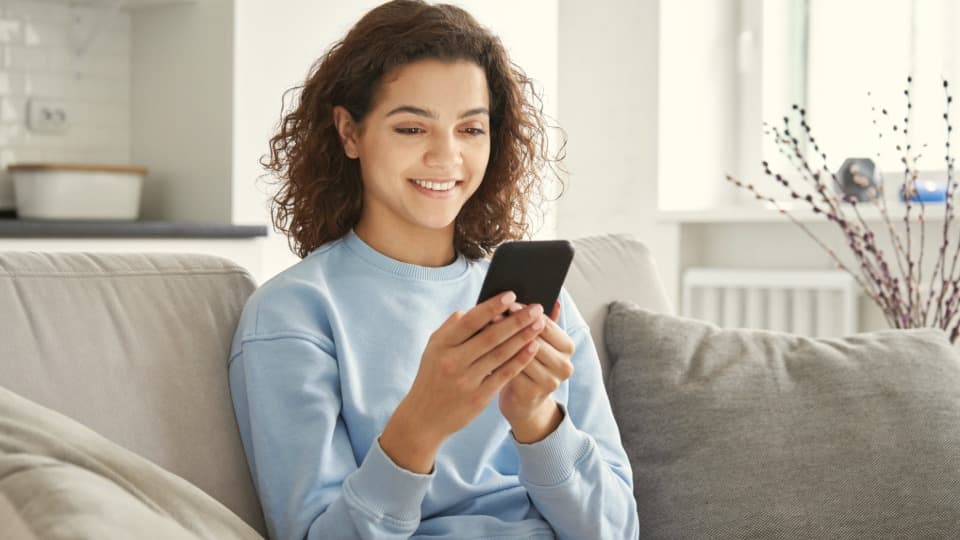 A young woman relaxes on a couch and uses her smartphone for the digital volume estimation or online video consultation for her move with DACHSER & KOLB.