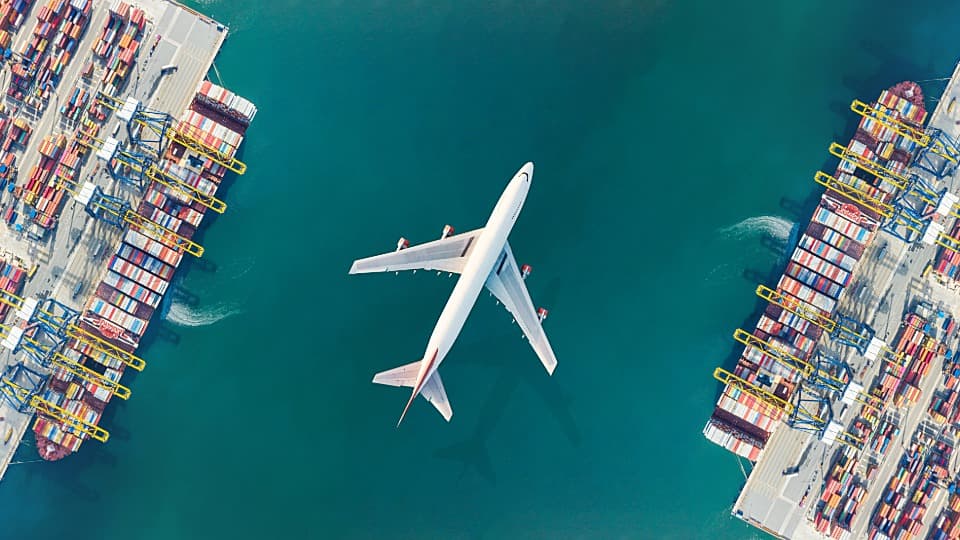 Eine Luftaufnahme zeigt ein Frachtflugzeug im Flug über zwei Containerschiffen im Hafen als Symbol für den Vergleich von Luftfracht und Seefracht beim Auslandsumzug.