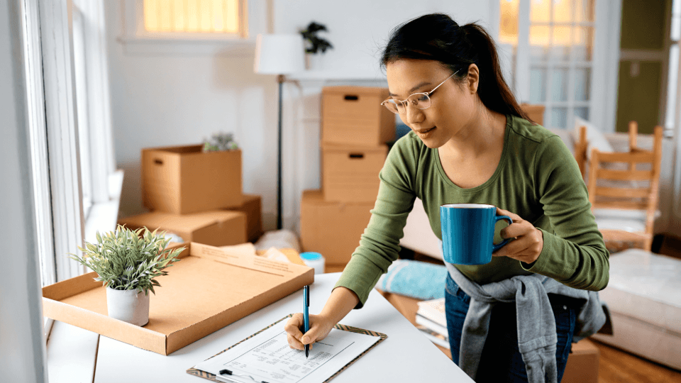 Woman filling out a high-value inventory list in an apartment with moving boxes, holding a blue mug.