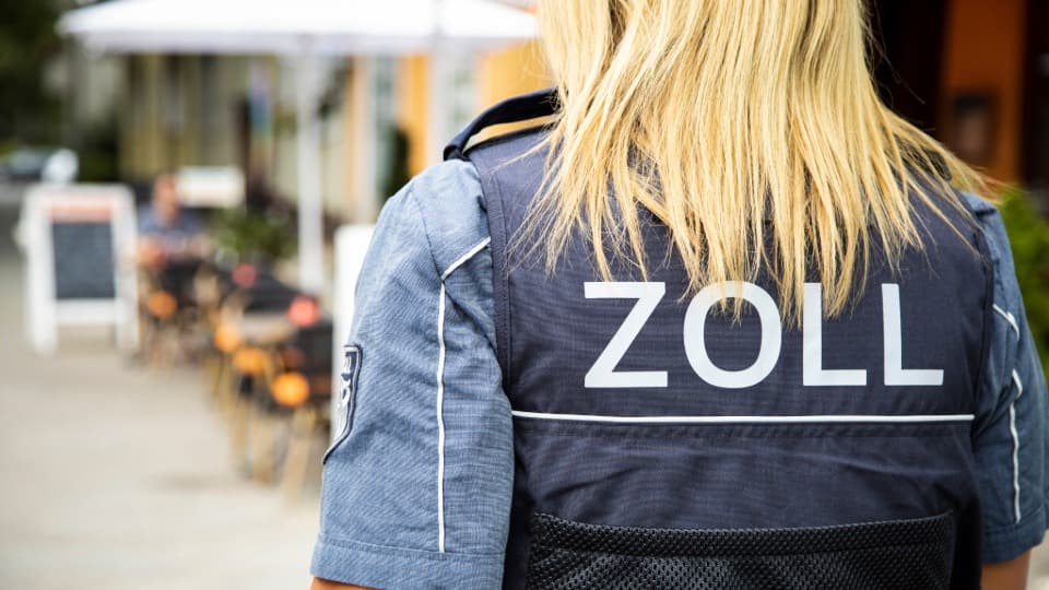Rear view of a customs officer wearing a uniform labeled “ZOLL” in a public area